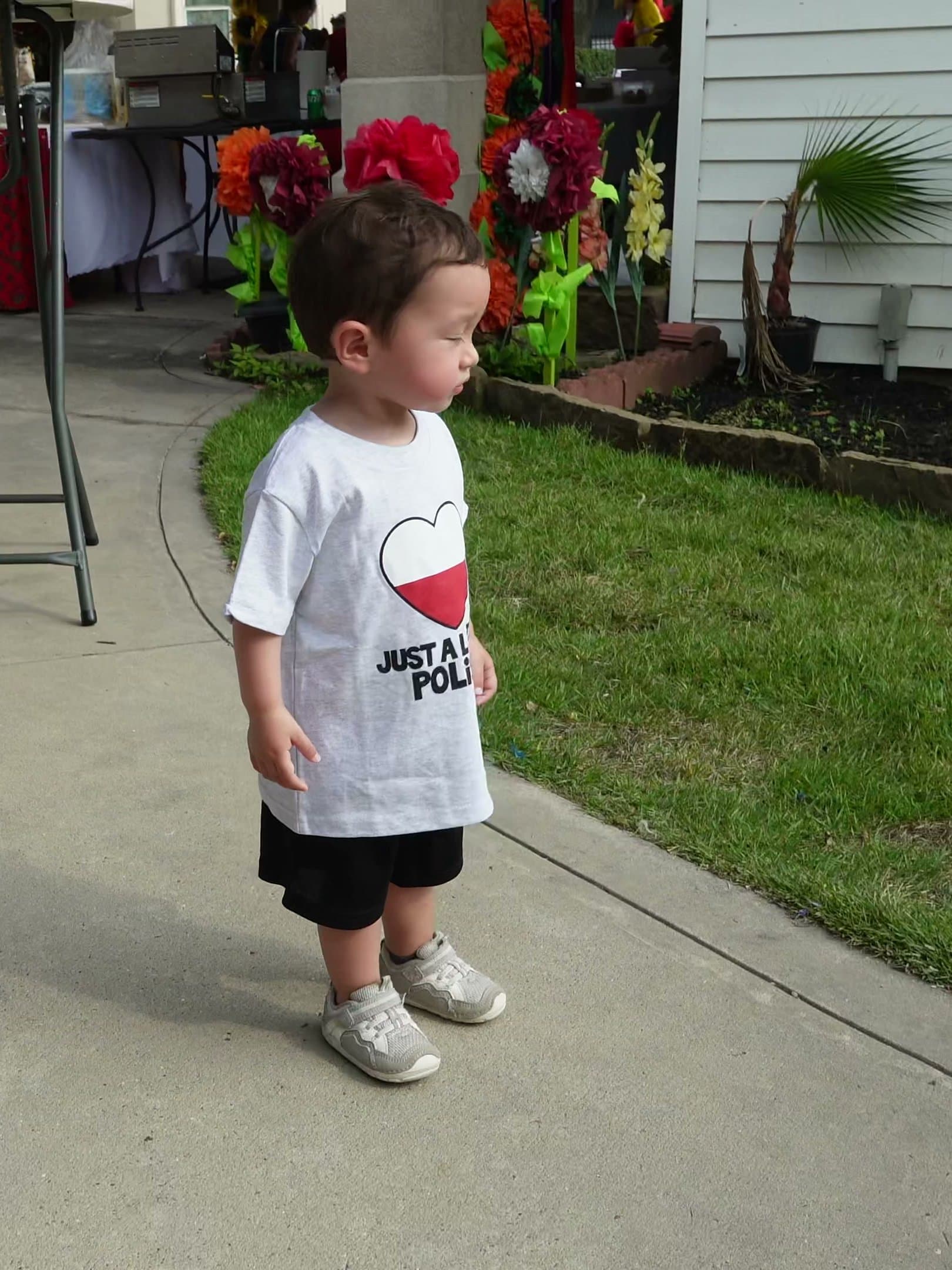 A toddler in a Polish-flag T-shirt standing on the path beside the festival's wall of oversized paper flowers.