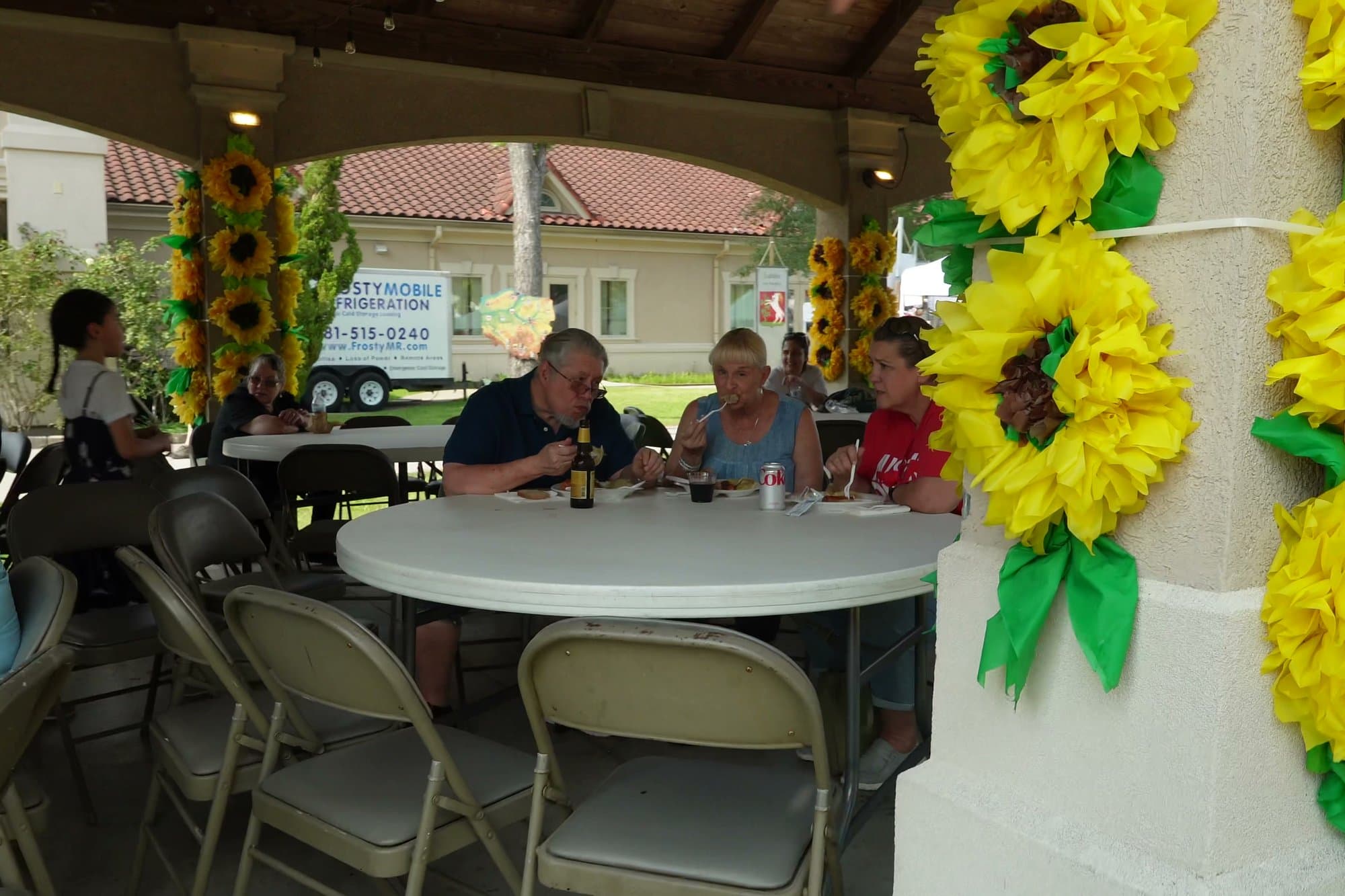 Festival-goers sitting at round tables under the open-sided pavilion, its posts wrapped in oversized paper sunflowers.