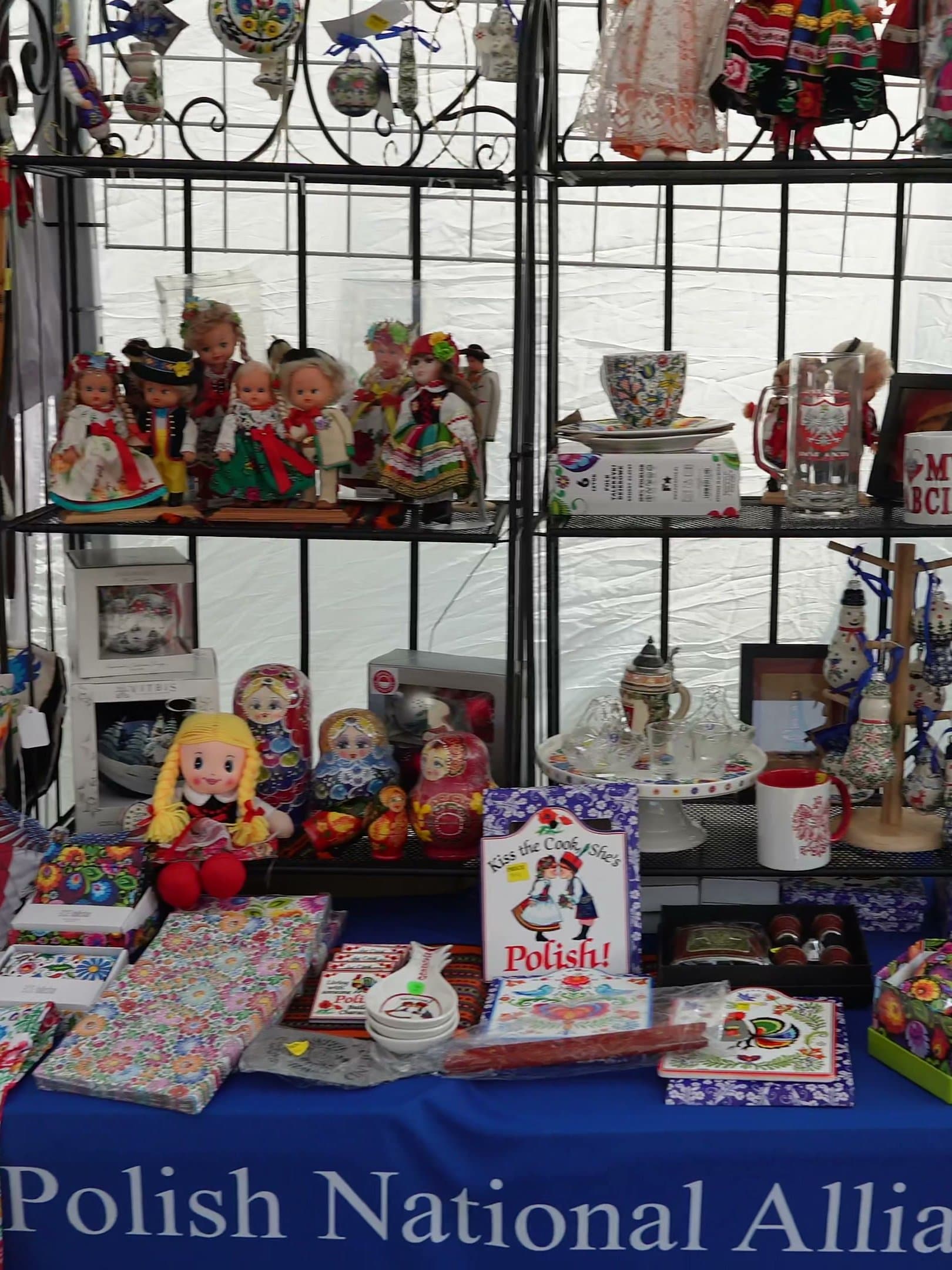 The Polish National Alliance stall, its shelves filled with folk dolls in regional dress, painted ceramics and books, under a hanging rack of embroidered blouses.