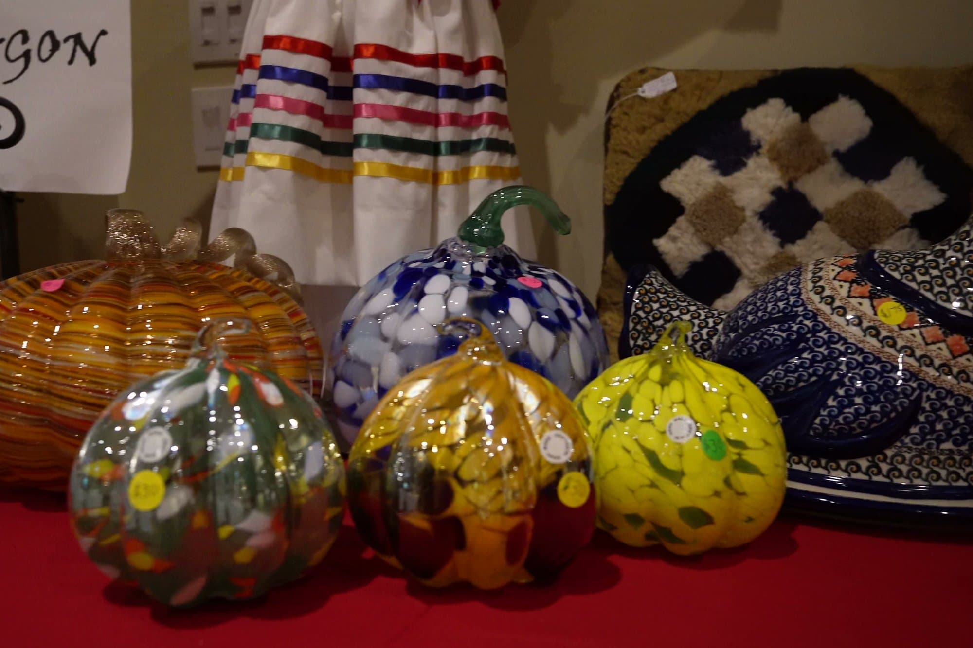 Hand-blown glass pumpkins in amber, cobalt and white standing on a red cloth beside a display of folk-patterned ceramics.