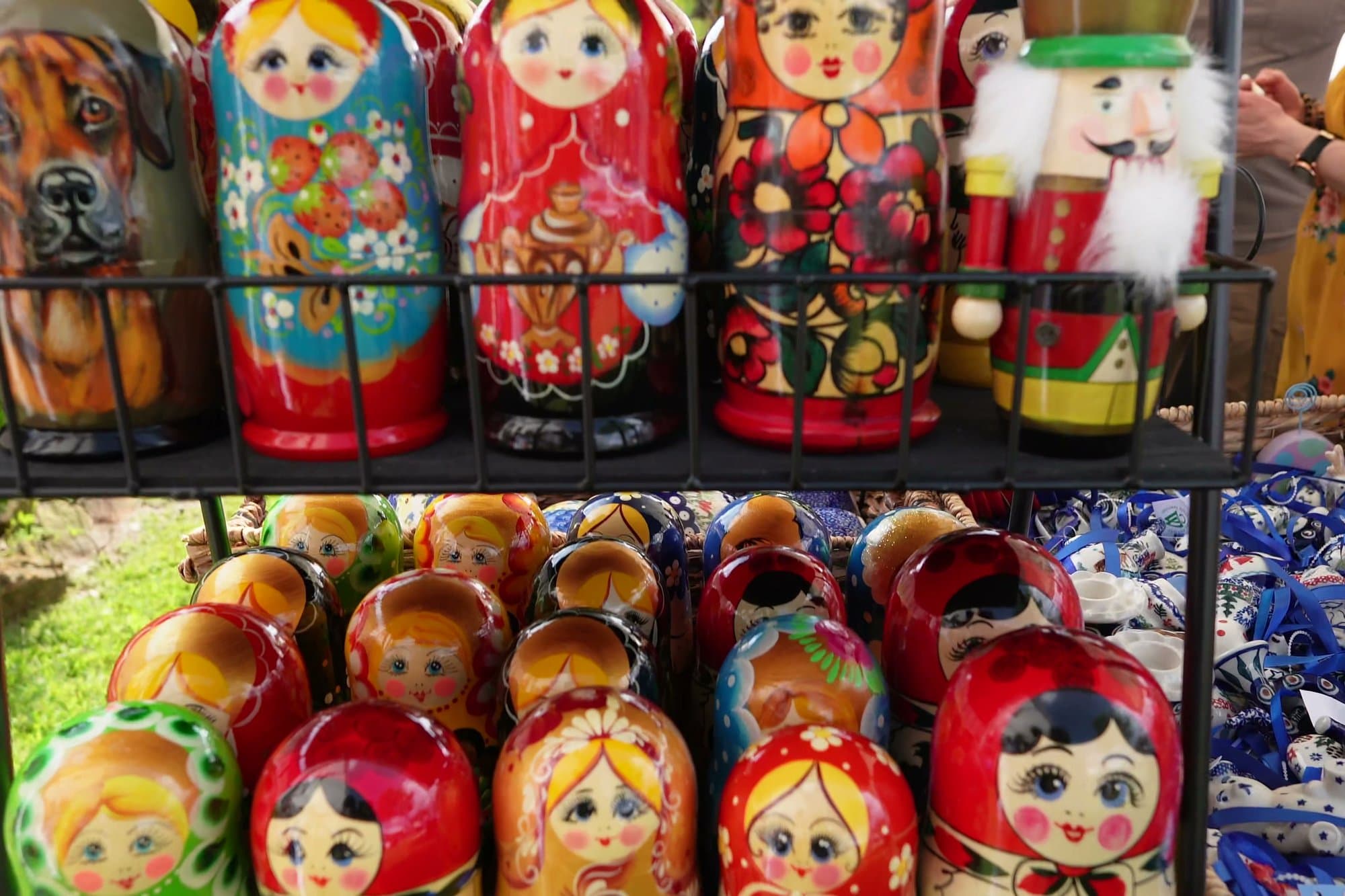 A market rack packed with painted matryoshka nesting dolls in rows of red, blue and gold, with a wooden nutcracker figure standing among them.