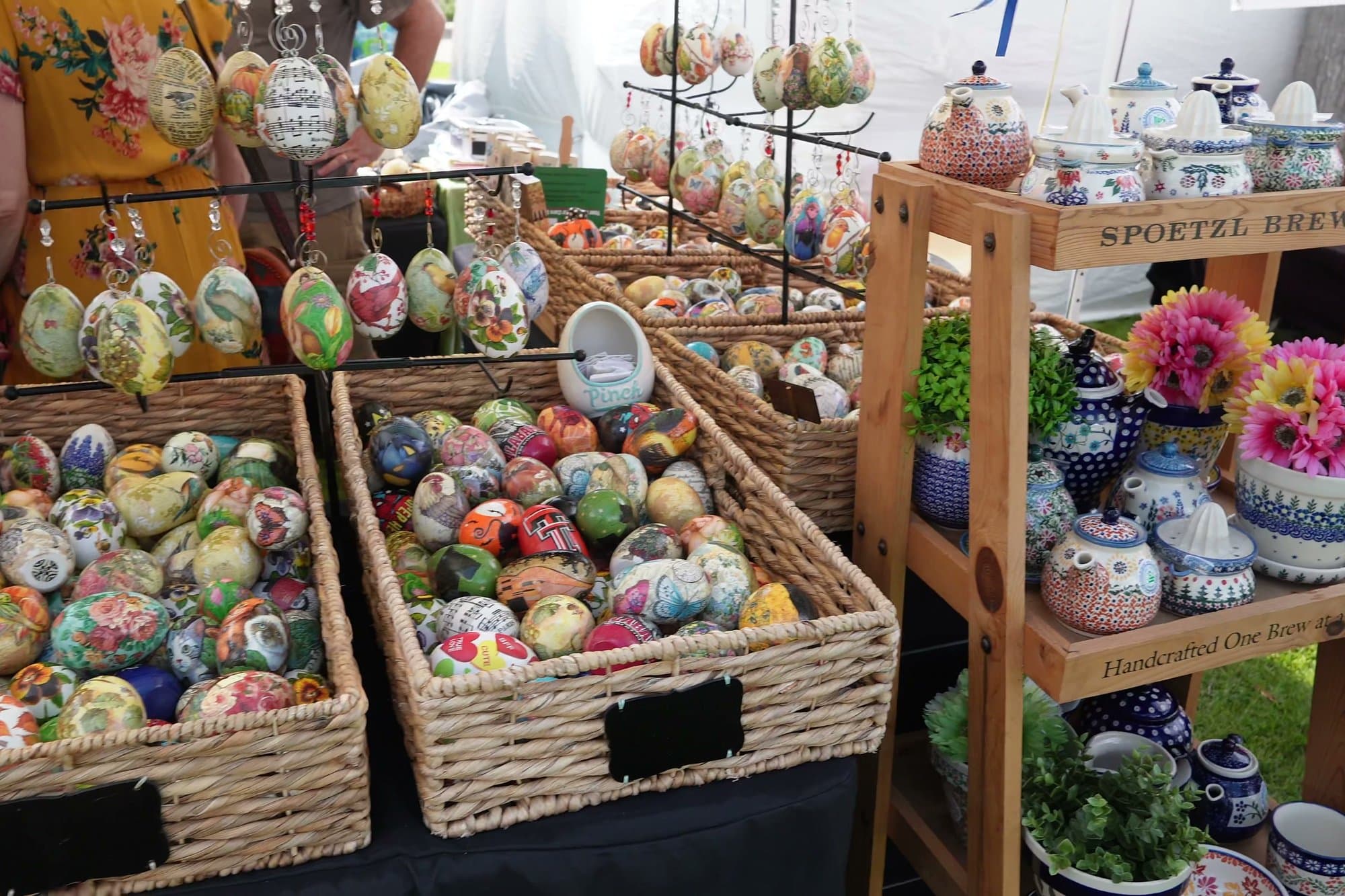 Wicker baskets heaped with hand-painted pisanki eggs in every colour, stacked on a market stall beside painted ceramics and potted flowers.