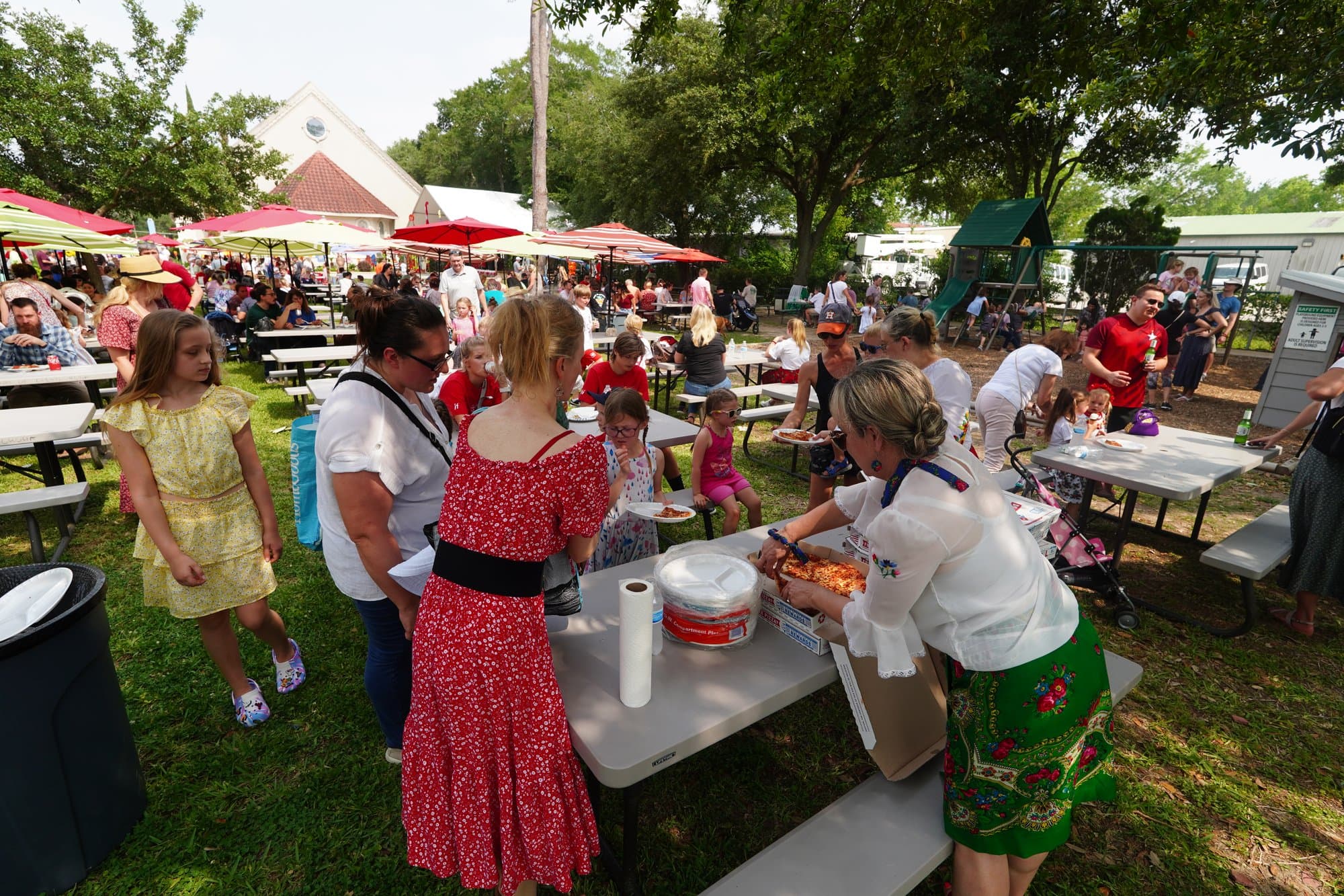 A wide view of the festival grounds: families at long picnic tables under shade trees and red umbrellas, volunteers in red and green folk skirts serving from a table, and a girl in a yellow dress in the foreground.