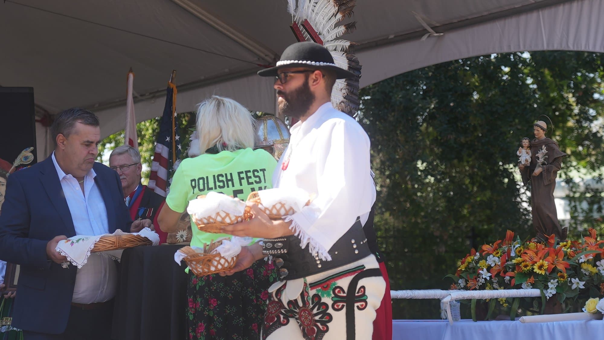 A man in Góral highlander dress — black felt hat, white trousers with embroidered appliqué and a wide leather belt — holding the decorated harvest loaf on the stage, with a volunteer in a green Polish Fest crew shirt beside him.