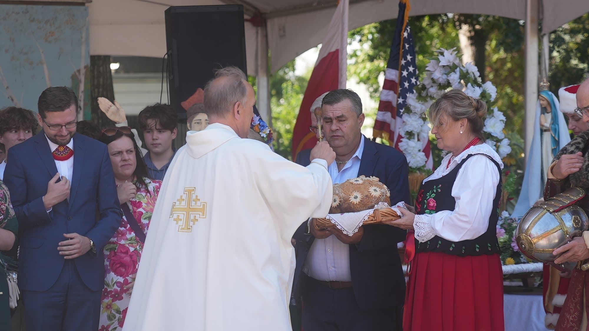 A priest in white vestments with a gold cross raising his hand to bless the harvest loaf as a couple hold it out to him. Behind them officials stand with hands over their hearts among flags and white floral decoration.