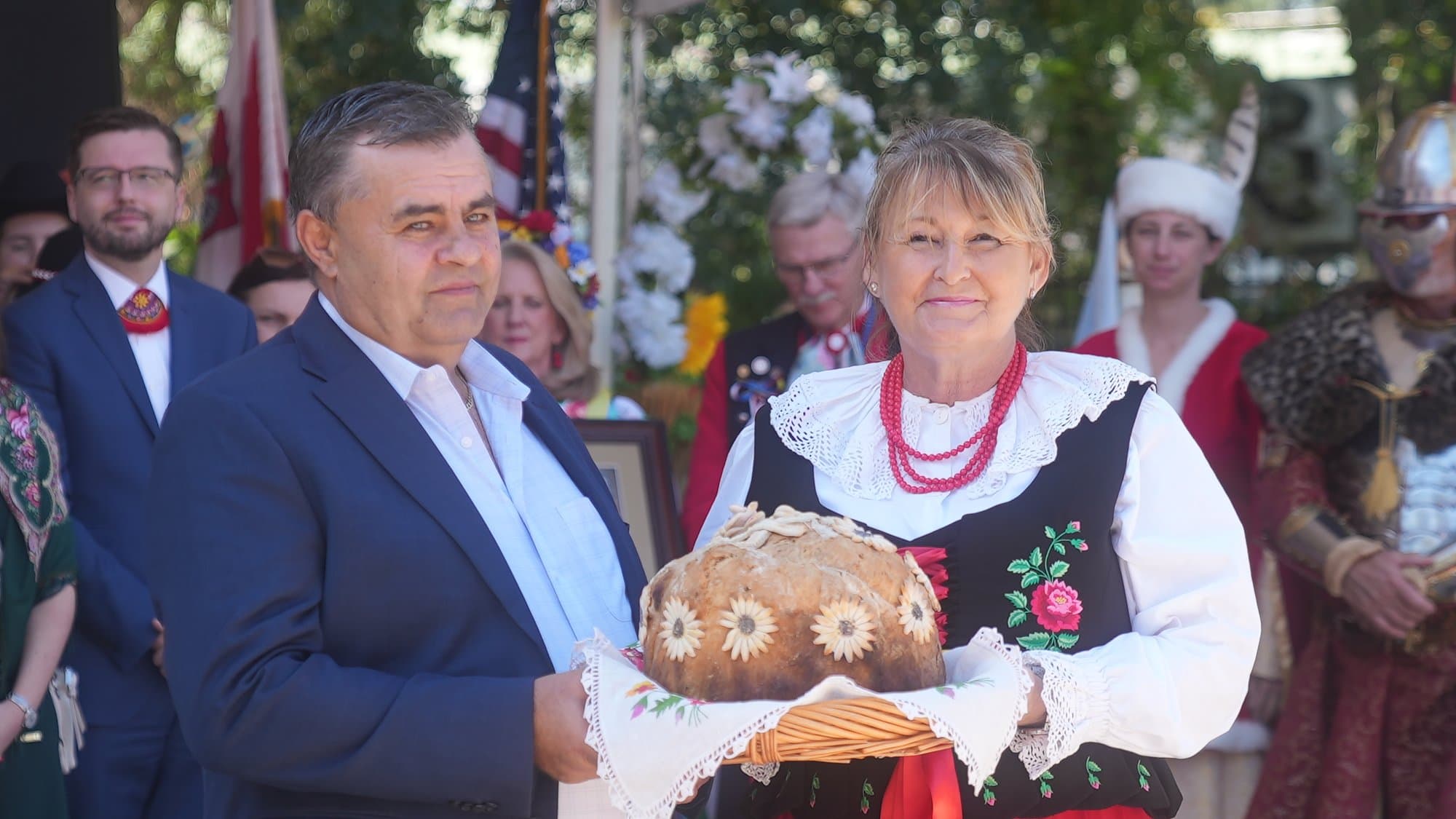 A woman in Kraków folk dress — black embroidered vest, white blouse and red coral beads — and a man in a navy suit together holding a large round harvest loaf, its crust decorated with baked daisy shapes, resting on an embroidered white cloth. Polish and American flags stand behind them.