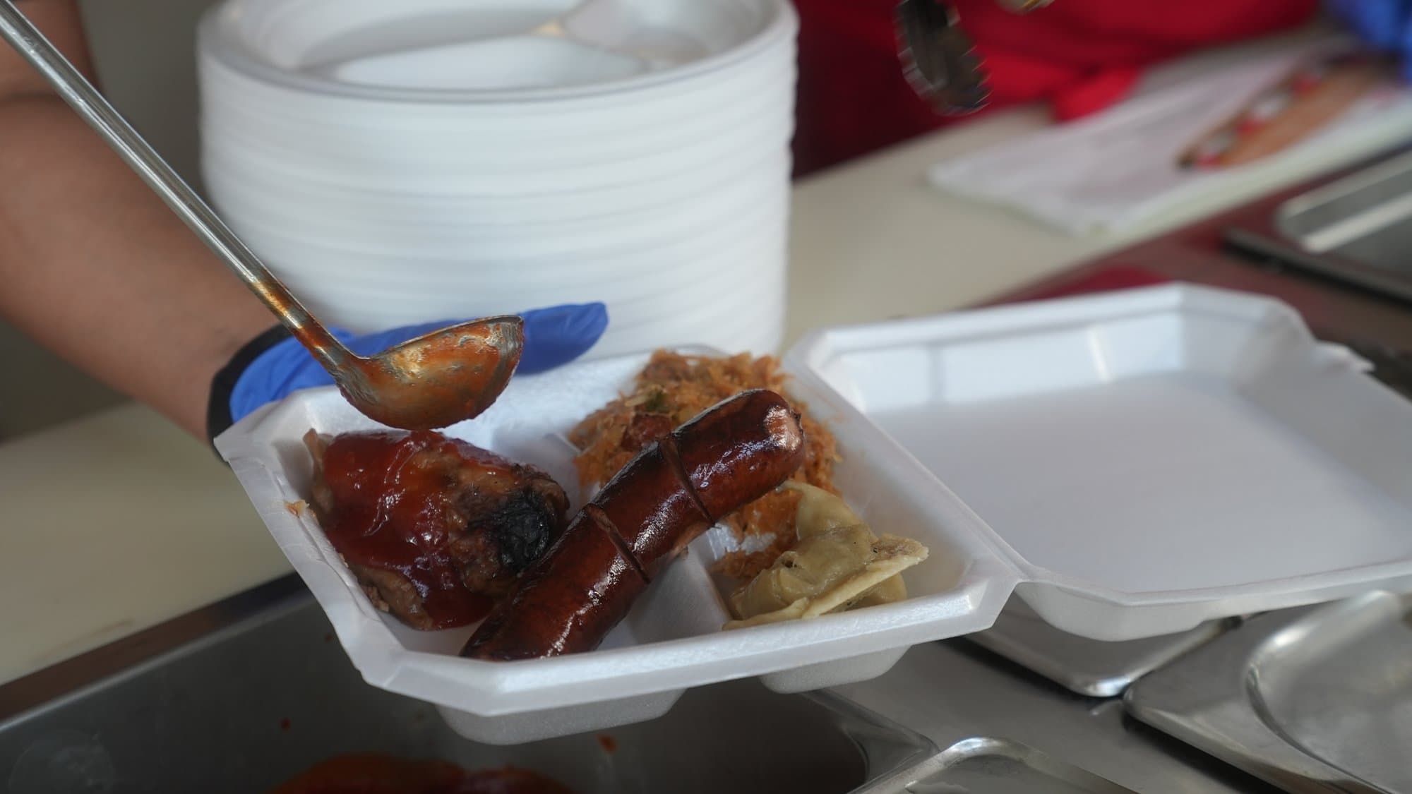 A gloved hand ladling sauce over grilled kiełbasa in a white takeaway box, with a tall stack of plates waiting behind.