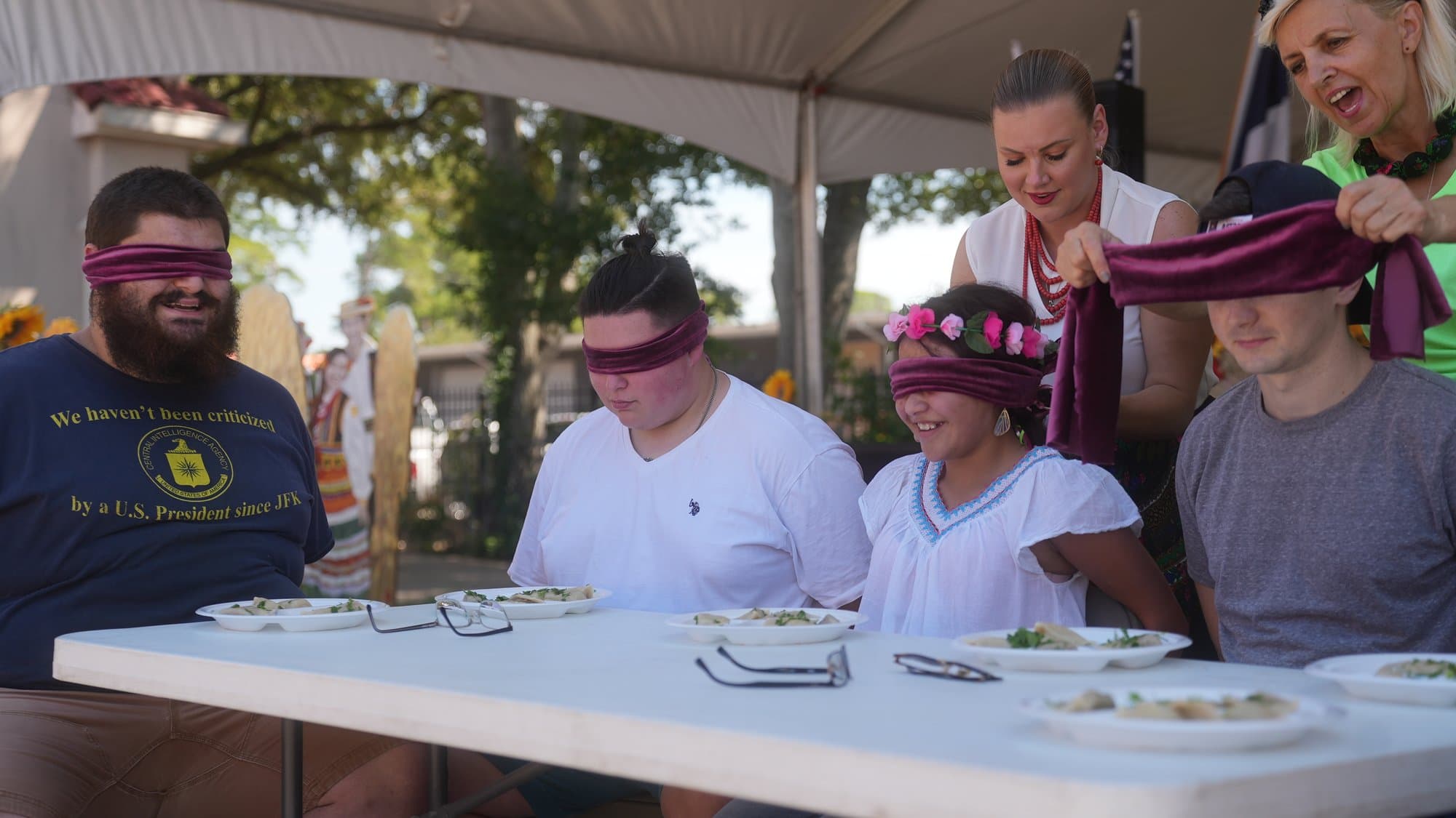 Four contestants sit blindfolded in maroon cloth at a long table set with plates of pierogi, while a host in a white blouse and red beads ties a blindfold on a young woman wearing a pink flower crown.