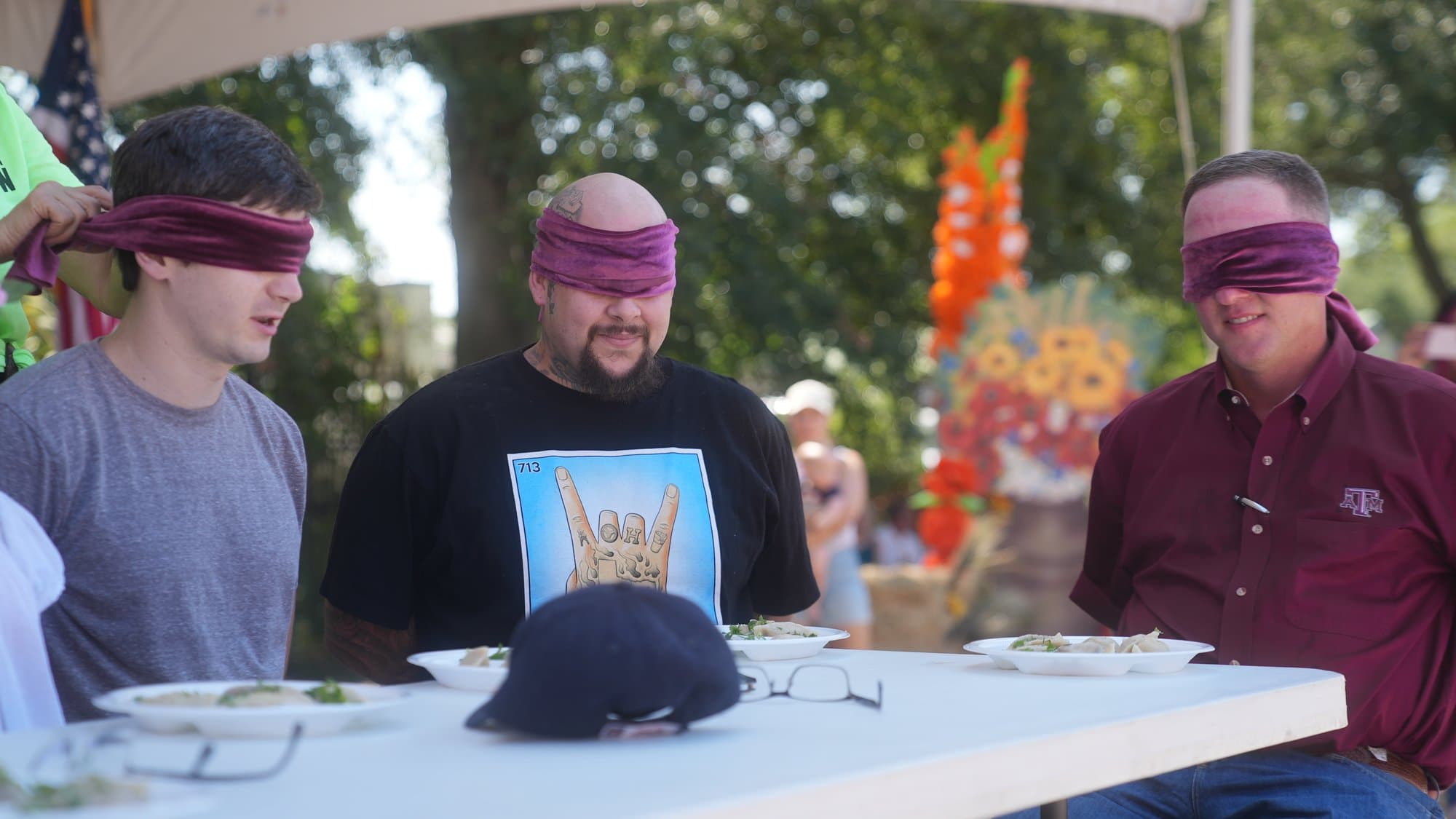 Three blindfolded men grinning at a white table, each with a plate of pierogi in front of them and their glasses folded on the tablecloth, a display of autumn flowers behind.