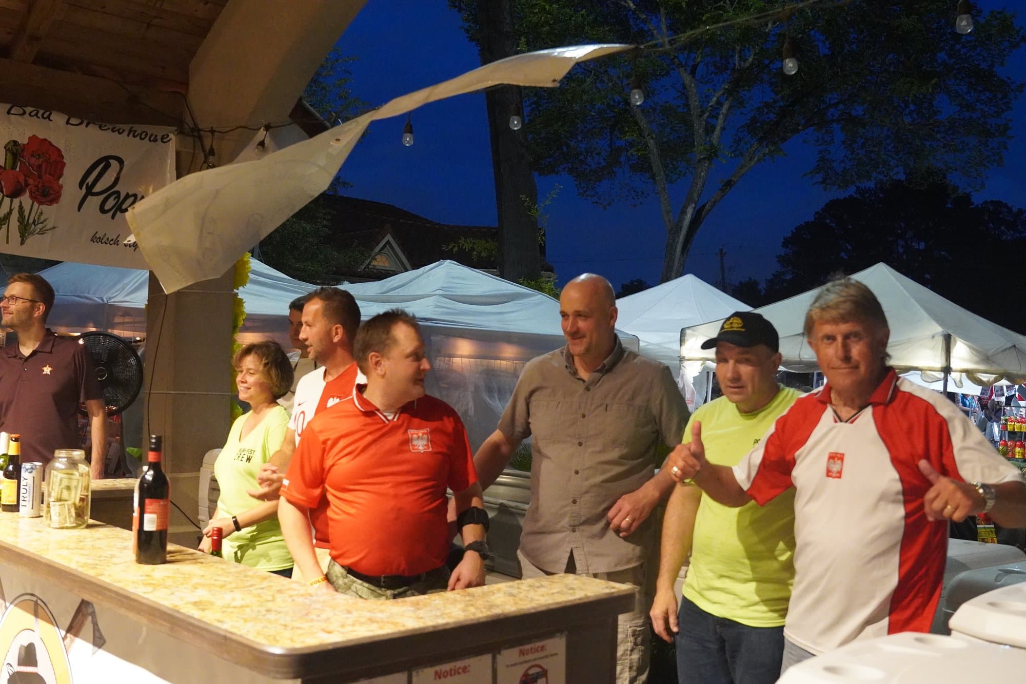 Volunteers laughing together behind the beer counter at dusk, wine and cans on the bar, marquees and a deep blue sky behind them.