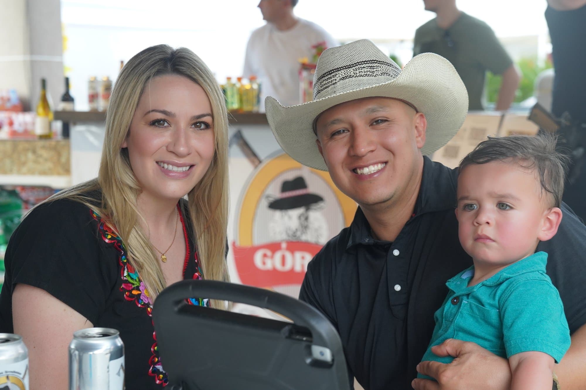 A couple with their toddler at the Góral bar, the father in a straw cowboy hat, the mother in black with floral embroidery, the festival's highlander logo on the counter behind them.