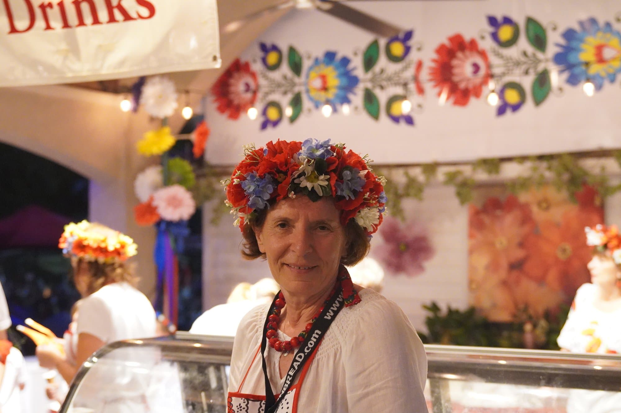 A woman in a white embroidered blouse, red coral beads and a full crown of flowers standing behind the serving counter, in front of a wall painted with bold folk flowers.