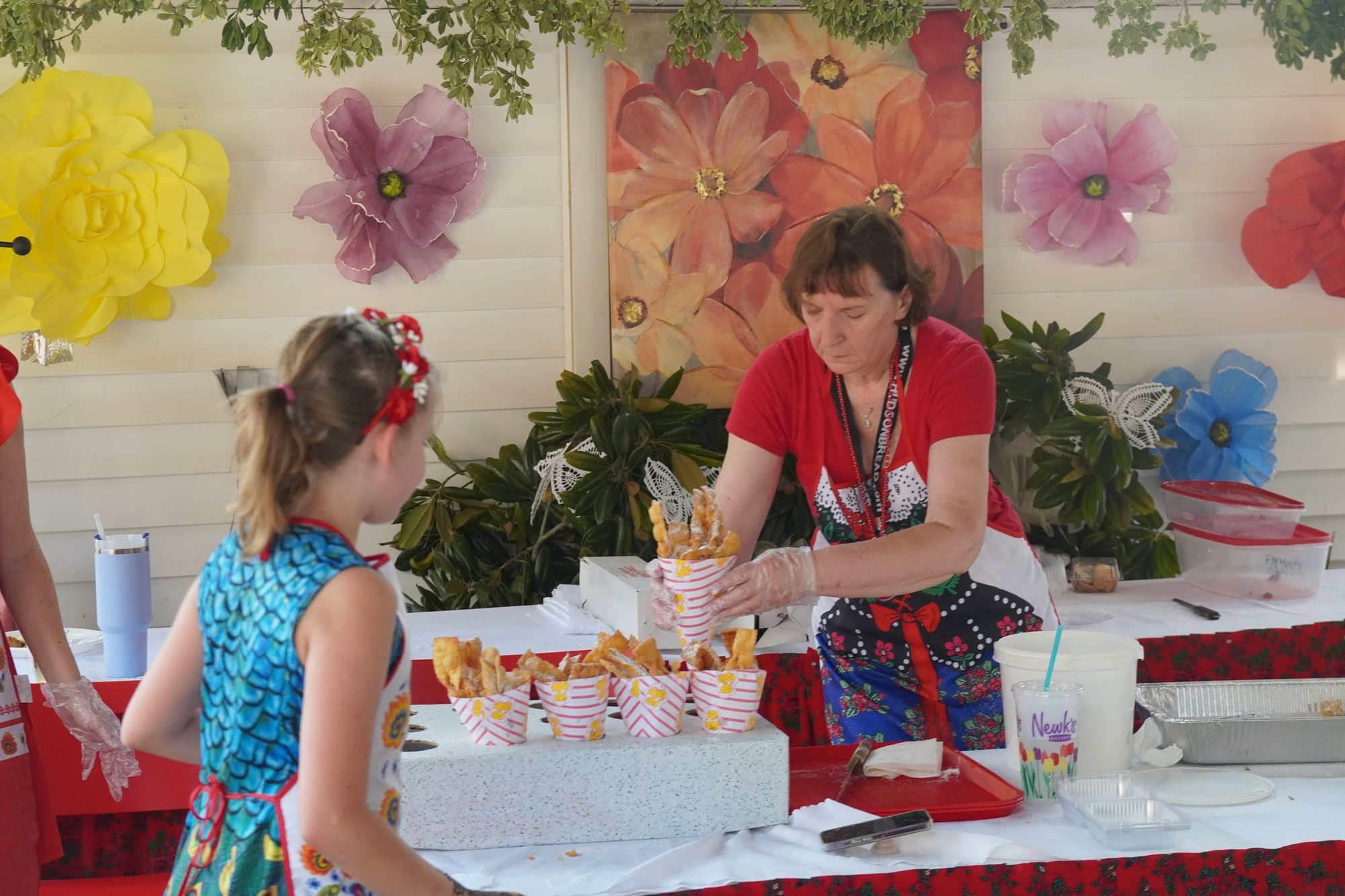 A girl in a bright blue floral folk dress and a red flower crown being served at the dessert table, where cartons of sugared pastry are lined up beneath a wall of oversized paper flowers.