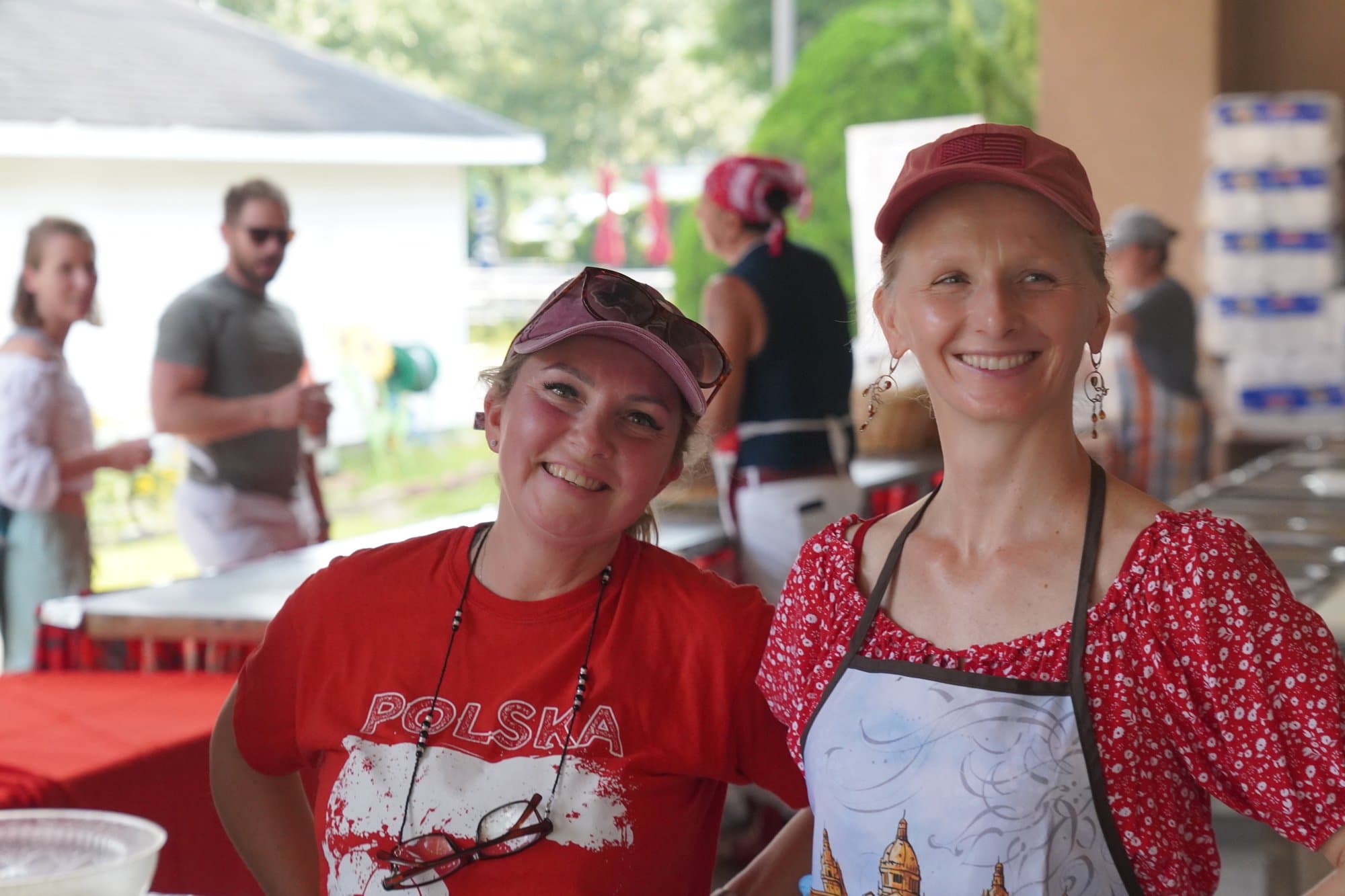Two volunteers smiling over the serving line, one in a red Polska shirt and visor, the other in a red floral apron.