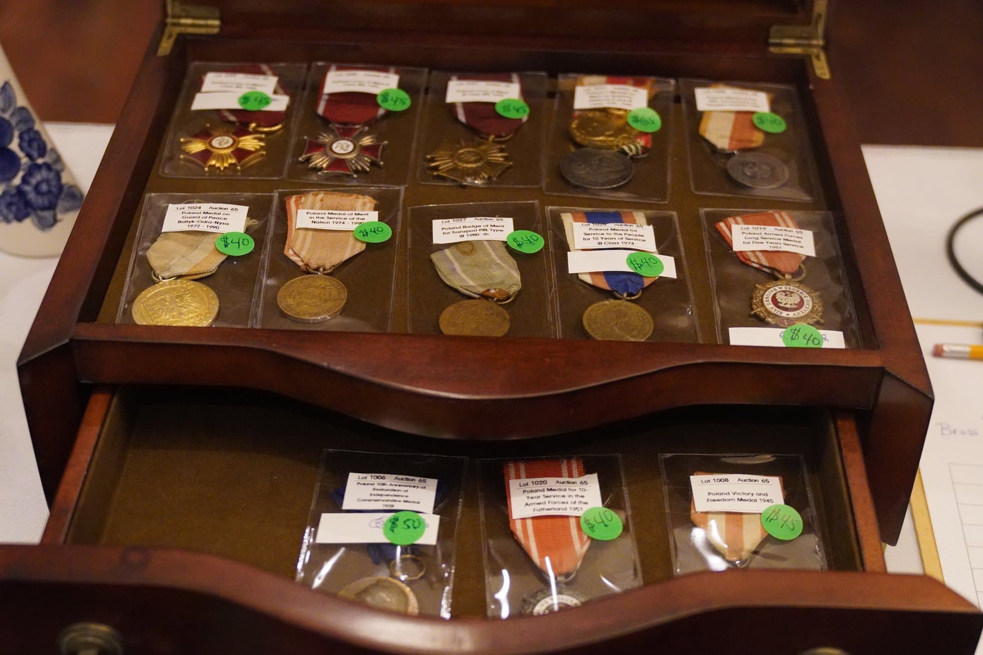A glass-topped wooden case displaying rows of commemorative medals on ribbons, each with a small handwritten tag.